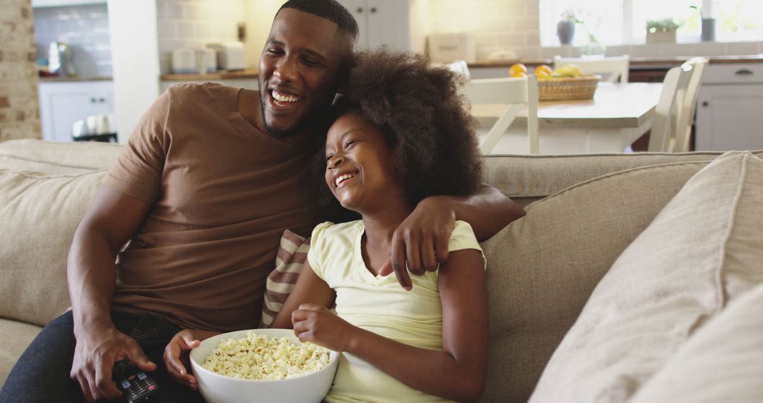 African american girl and her dad on couch eating popcorn and laughing at tv - Free Images, Stock Photos and Pictures on Pikwizard.com