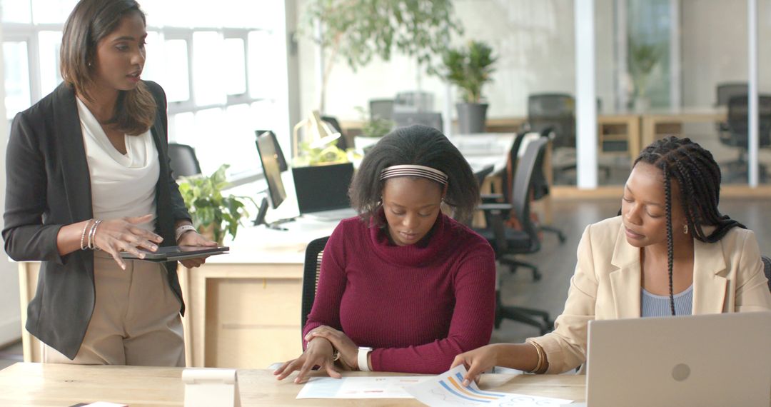 Diverse female team collaborating over charts with tablet and laptop in modern office