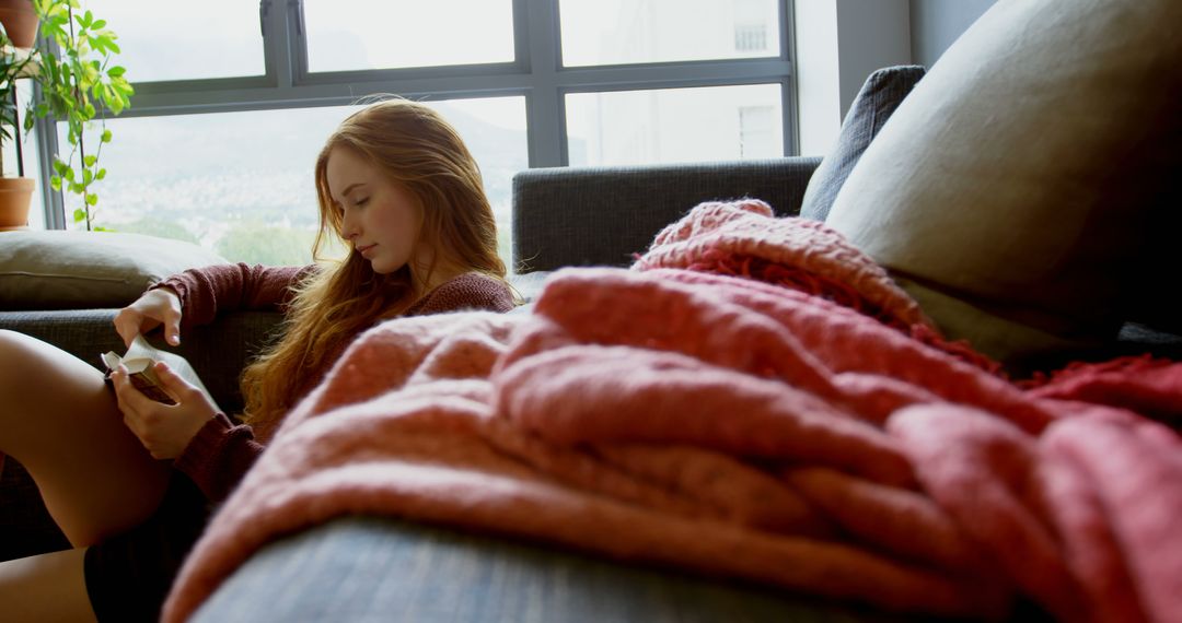 Young woman relaxing on couch reading a book by the window - Free Images, Stock Photos and Pictures on Pikwizard.com