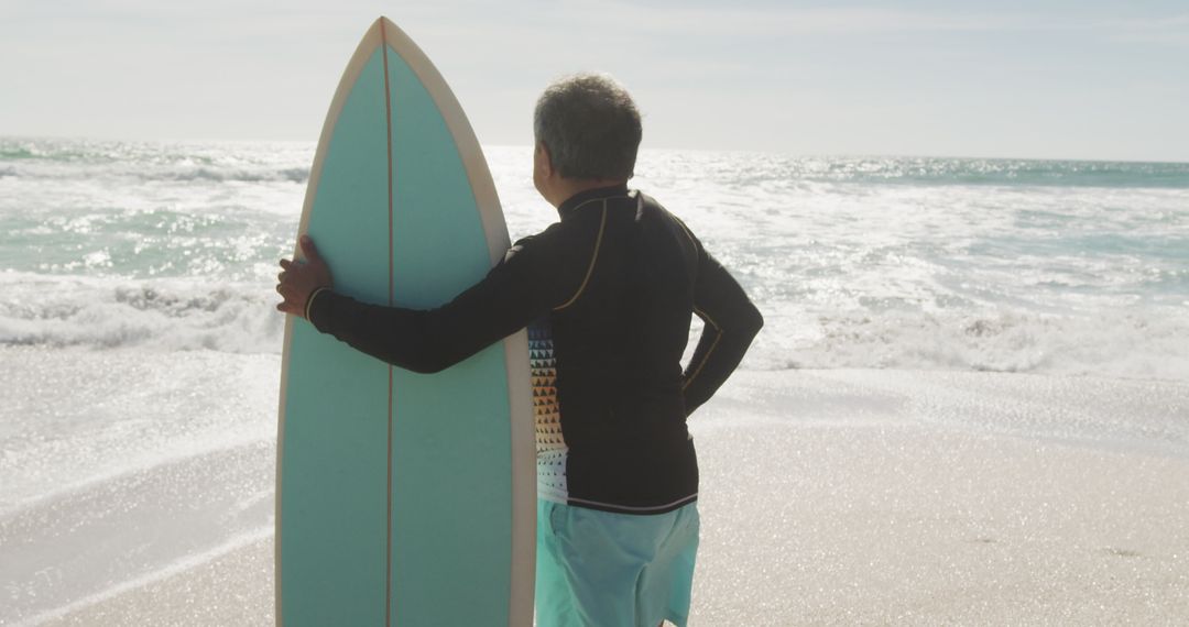 Senior Man Contemplating Ocean with Surfboard on Sunny Beach