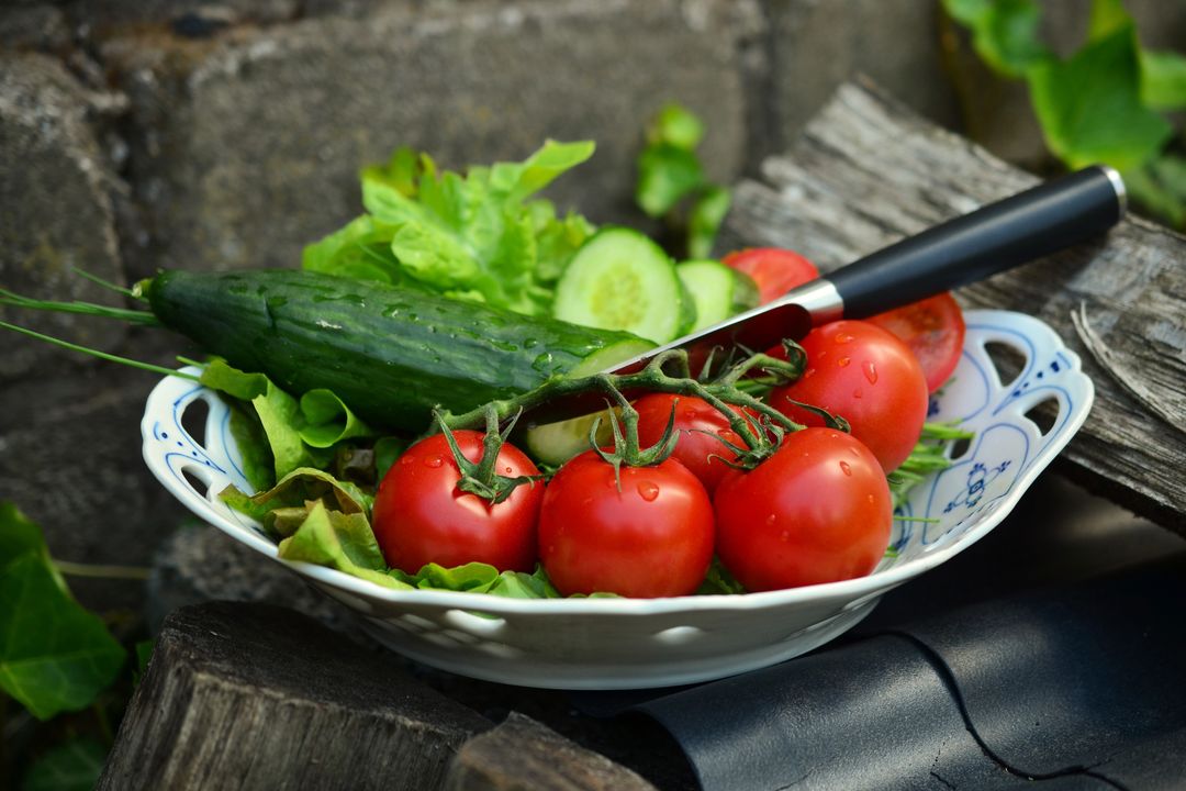 Plump vine tomatoes and cucumber resting on vintage bowl with leafy salad and knife