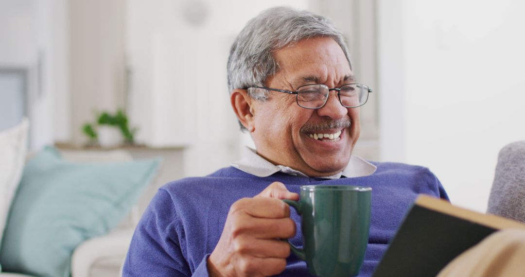 Image of happy senior biracial man drinking coffee, reading book at home and laughing - Free Images, Stock Photos and Pictures on Pikwizard.com
