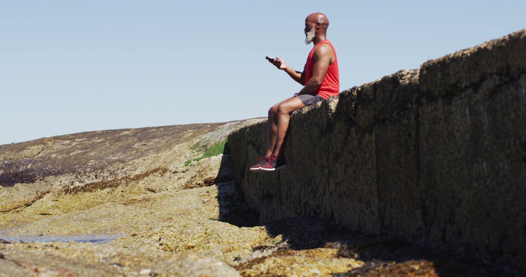 Elderly Man Relaxing on Coastal Wall with Smartphone on Sunny Day - Free Images, Stock Photos and Pictures on Pikwizard.com