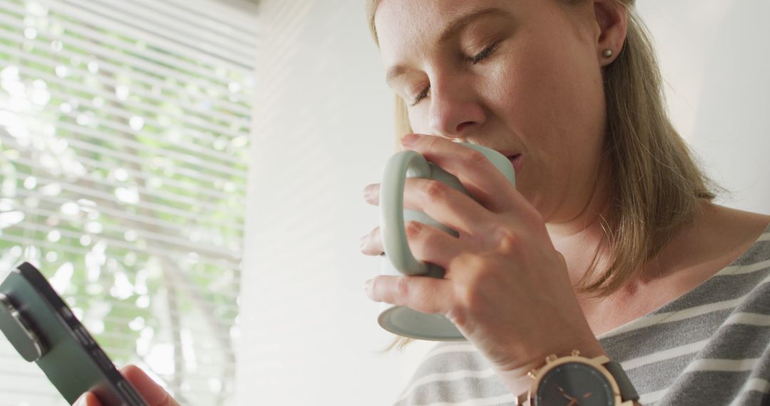 Woman drinking coffee while using smartphone near window with blinds - Free Images, Stock Photos and Pictures on Pikwizard.com