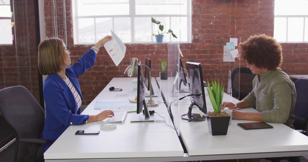 Woman showing chart while colleague types across clear divider in modern loft workspace