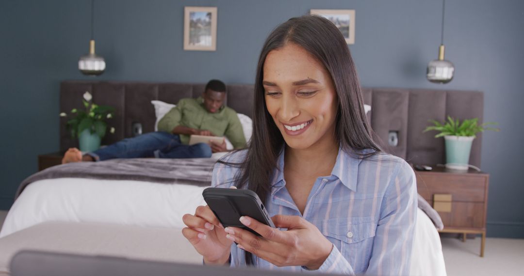 Smiling Woman Using Smartphone in Cozy Bedroom with Partner Relaxing - Free Images, Stock Photos and Pictures on Pikwizard.com