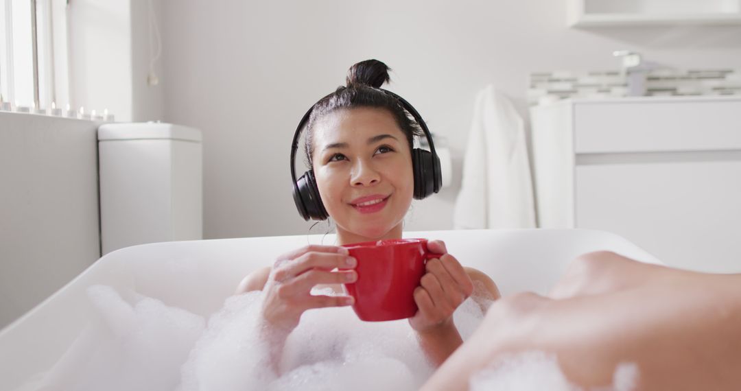 Image of smiling biracial woman with headphones and red cup of tea in bathtub in bubble bath - Free Images, Stock Photos and Pictures on Pikwizard.com