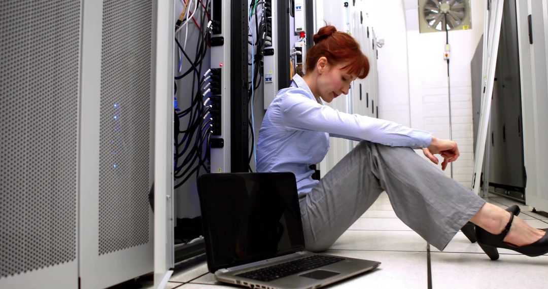 Sitting IT technician troubleshooting laptop between server racks in data center