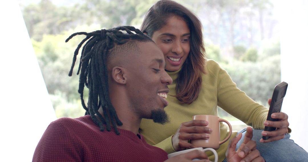 Couple sharing coffee and smartphone on couch, smiling and relaxing in cozy home