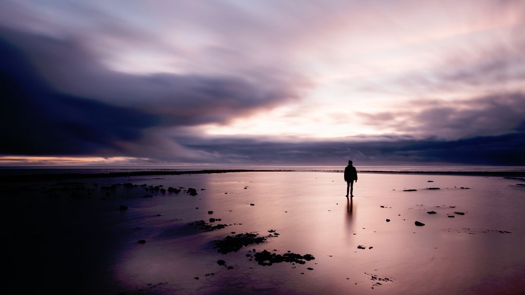 Solitary Figure Standing on Reflective Shore at Purple Sunset with Long-Exposure Clouds