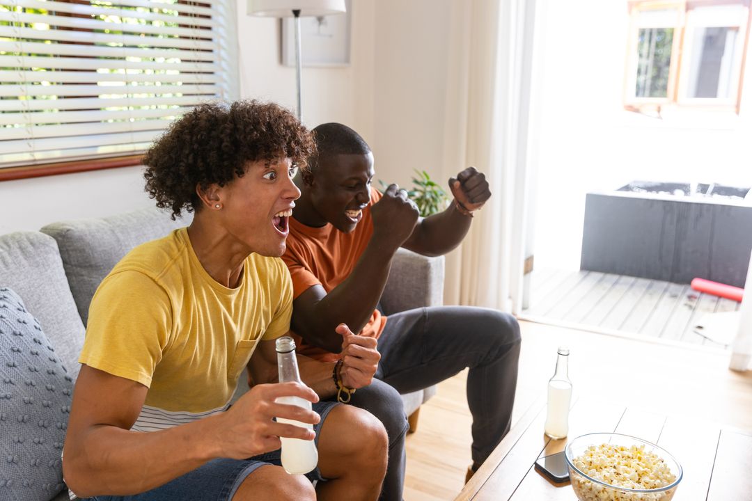 Young diverse friends cheering on sofa watching game holding drinks and popcorn