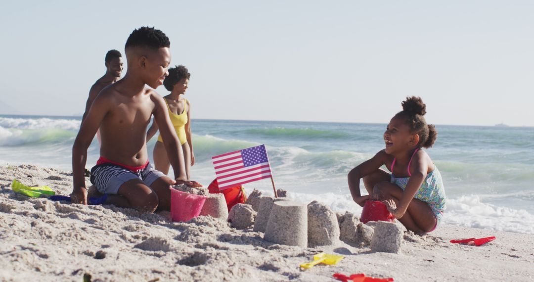 Smiling african american family building sandcastle with american flag on sunny beach - Free Images, Stock Photos and Pictures on Pikwizard.com