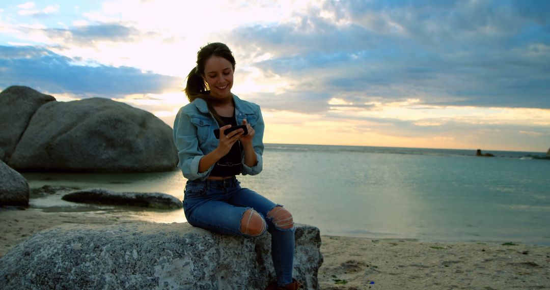 Woman Sitting on Rock by Seaside Beach Reading Message on Phone During Sunset - Free Images, Stock Photos and Pictures on Pikwizard.com