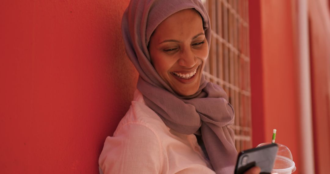 Muslim Woman Smiling and Using Smartphone While Holding Beverage - Free Images, Stock Photos and Pictures on Pikwizard.com