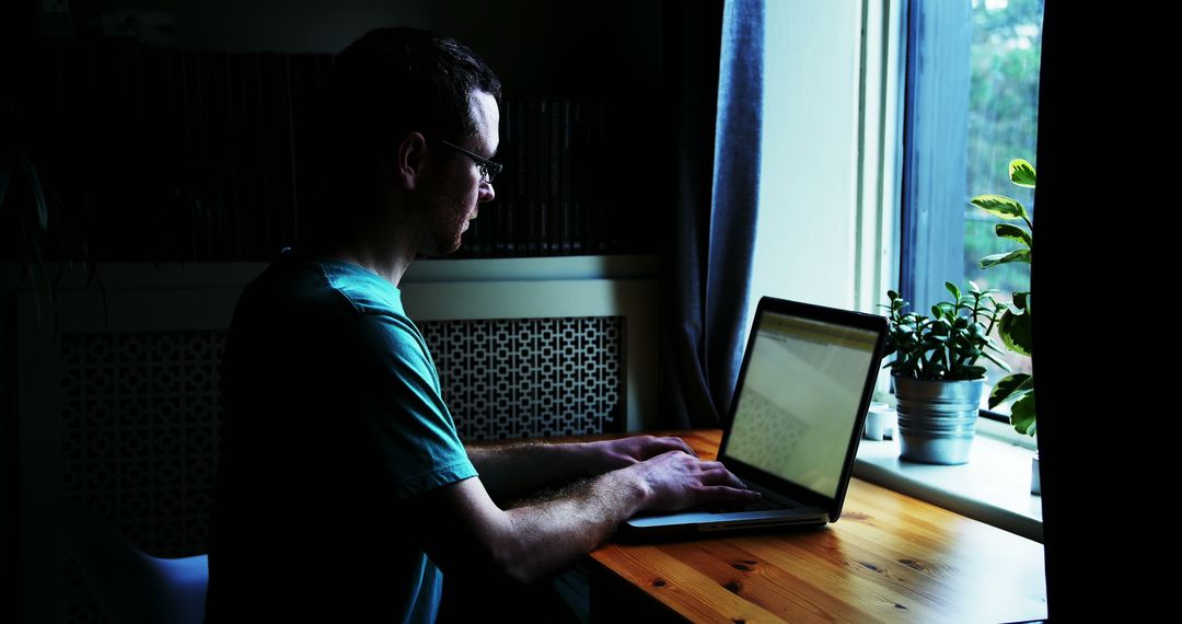 Young Man Working on Laptop by Window in Dimly Lit Room - Free Images, Stock Photos and Pictures on Pikwizard.com