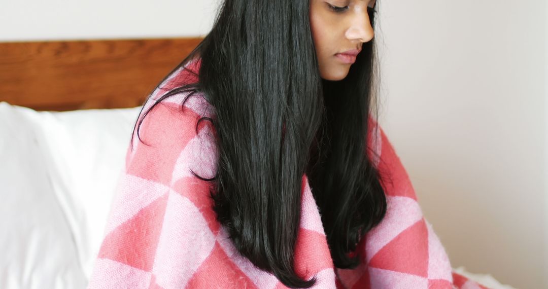 Sad biracial woman with long hair covered with blanket sitting on bed at home - Free Images, Stock Photos and Pictures on Pikwizard.com