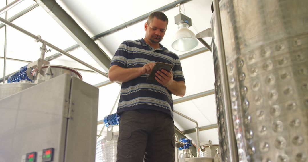 Man in brewery checking tablet amidst large metal equipment - Free Images, Stock Photos and Pictures on Pikwizard.com