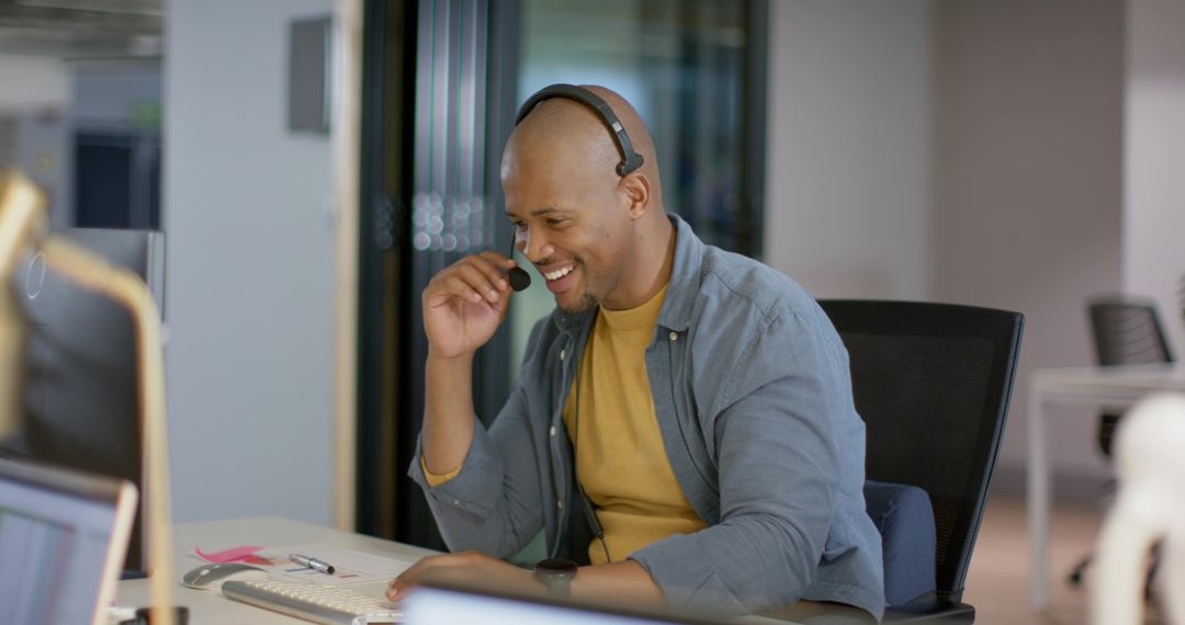 African American man smiling while using headset and typing at open office workstation