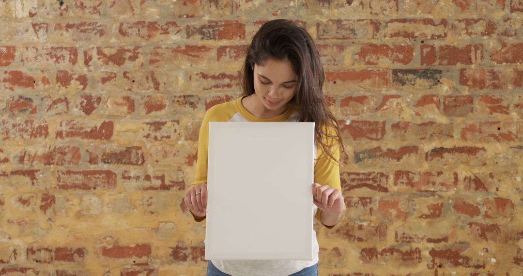 Portrait of happy biracial woman with long hair holding white board with copy space by brick wall - Free Images, Stock Photos and Pictures on Pikwizard.com