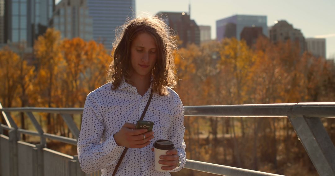 Young Man with Long Hair Looks at Phone on City Bridge During Autumn - Free Images, Stock Photos and Pictures on Pikwizard.com