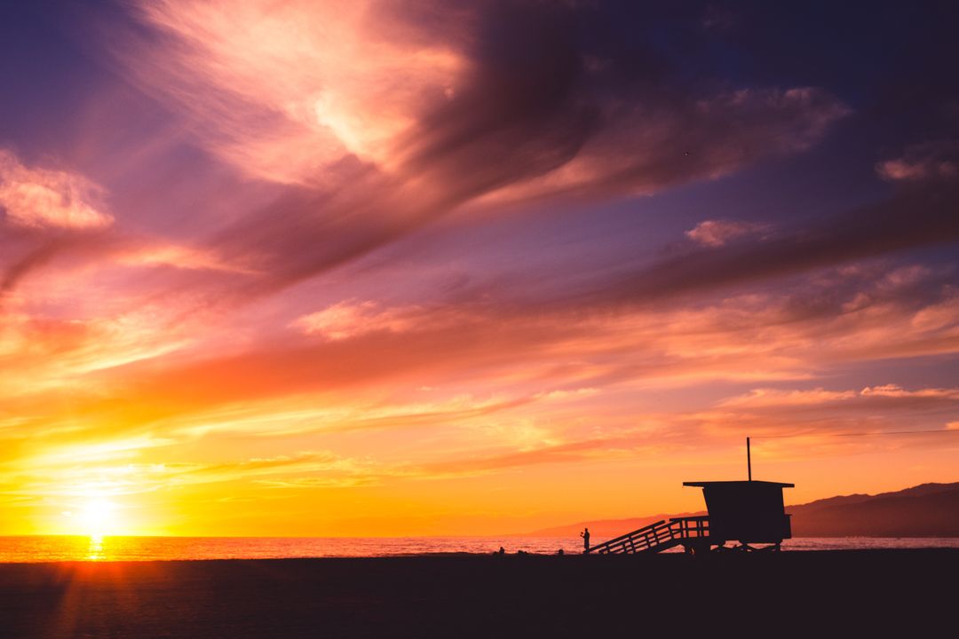 Sunset Sky Glowing Over Lifeguard Tower and Person Walking on Beach
