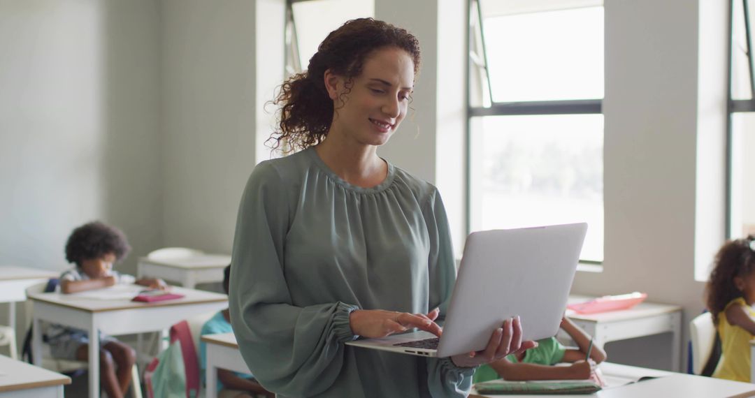 Smiling teacher teaching while using laptop in bright classroom with focused students