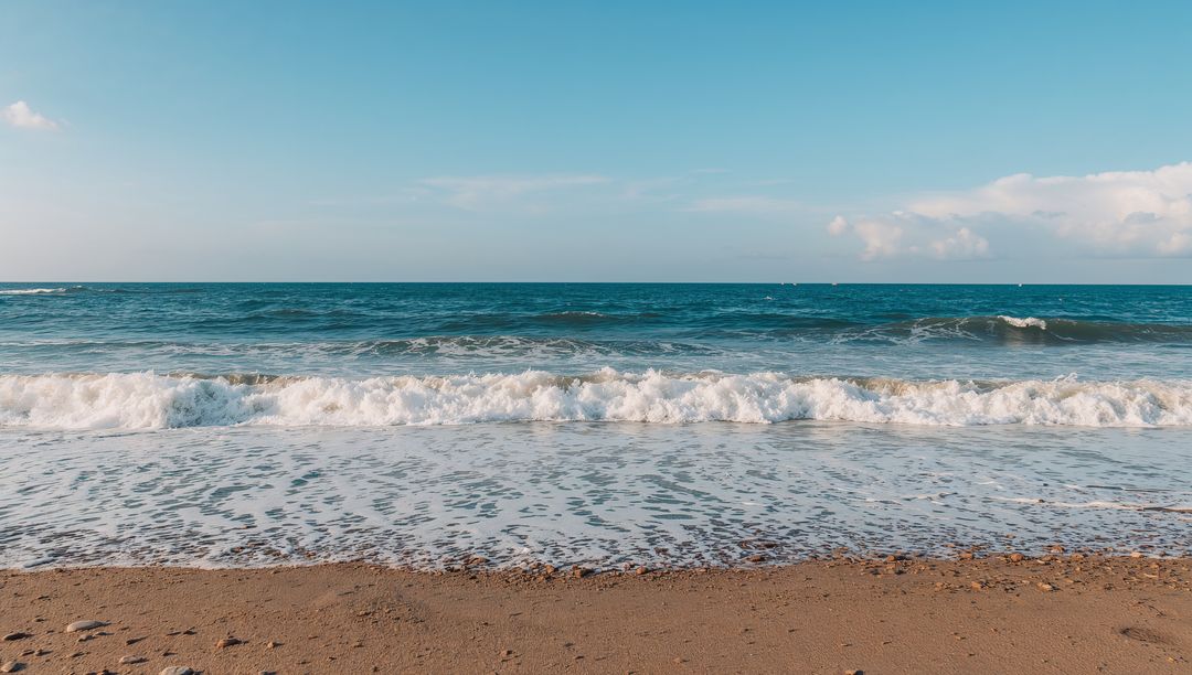 Sunlit breaking waves rolling onto sandy shore with white foam and pebbles