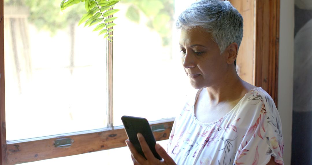 Senior Woman Using Smartphone by Sunny Window at Home