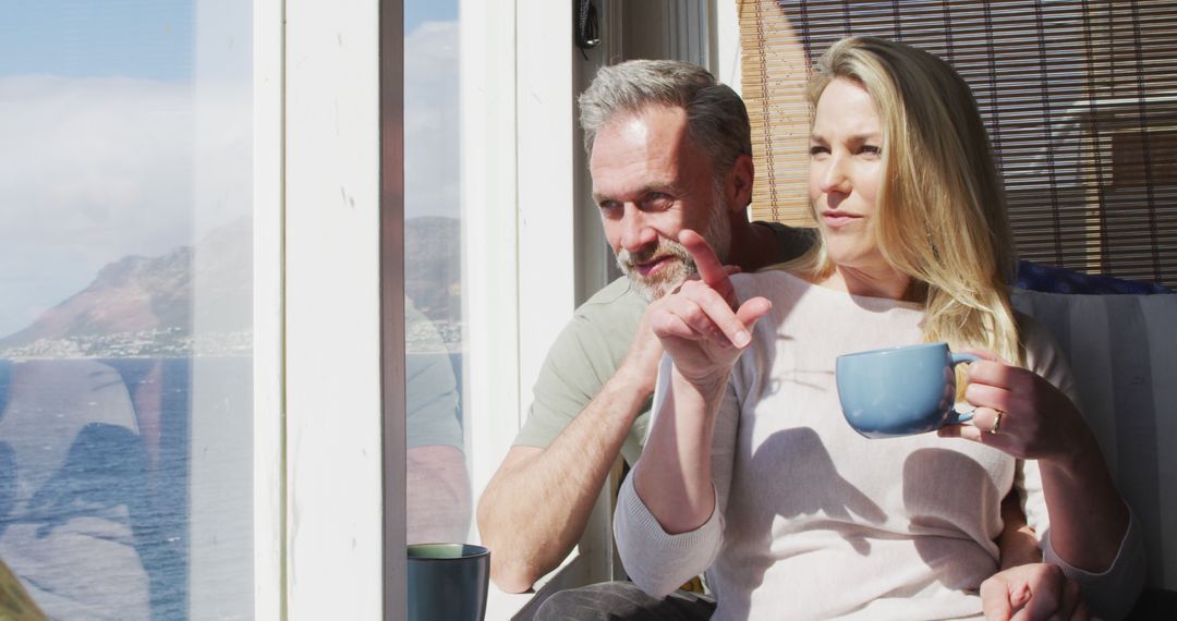 Happy Couple Enjoying Morning Coffee by Large Window with Ocean View - Free Images, Stock Photos and Pictures on Pikwizard.com