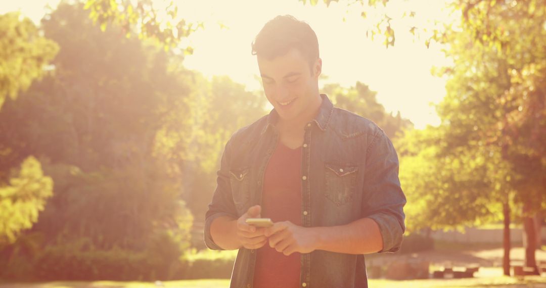 Young man smiling while using a smartphone outdoors under sunlight - Free Images, Stock Photos and Pictures on Pikwizard.com