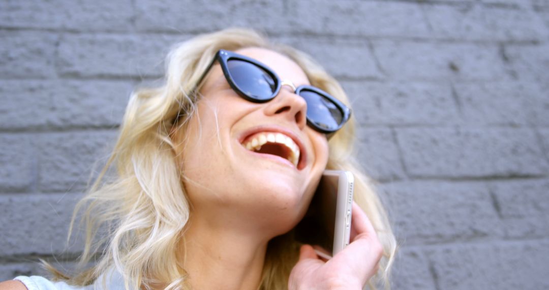 Blonde Woman Laughing While Talking on Smartphone Against Brick Wall - Free Images, Stock Photos and Pictures on Pikwizard.com