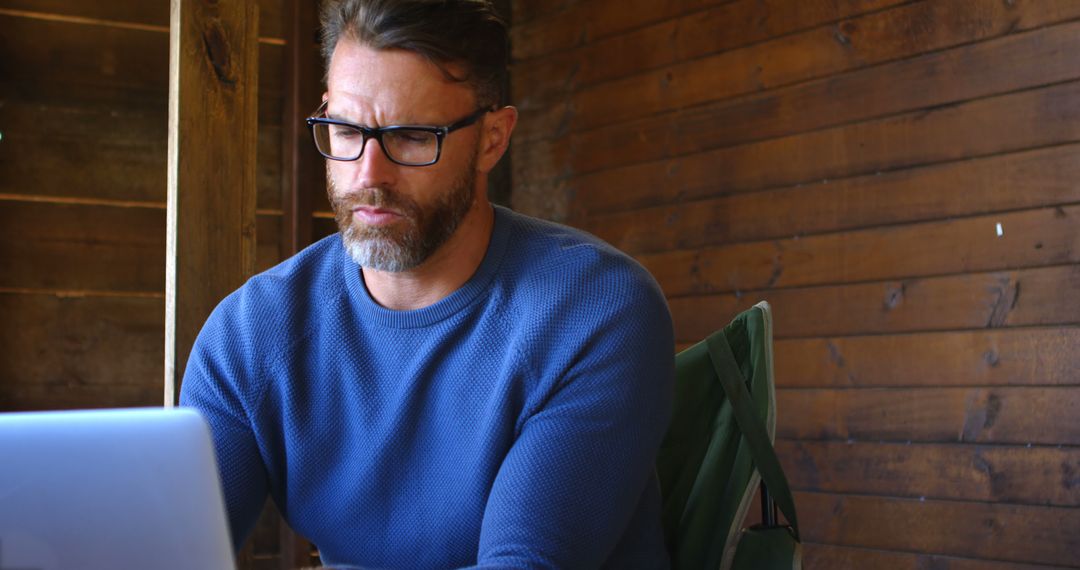 Bearded Man Wearing Glasses Working on Laptop in Rustic Wooden Cabin - Free Images, Stock Photos and Pictures on Pikwizard.com