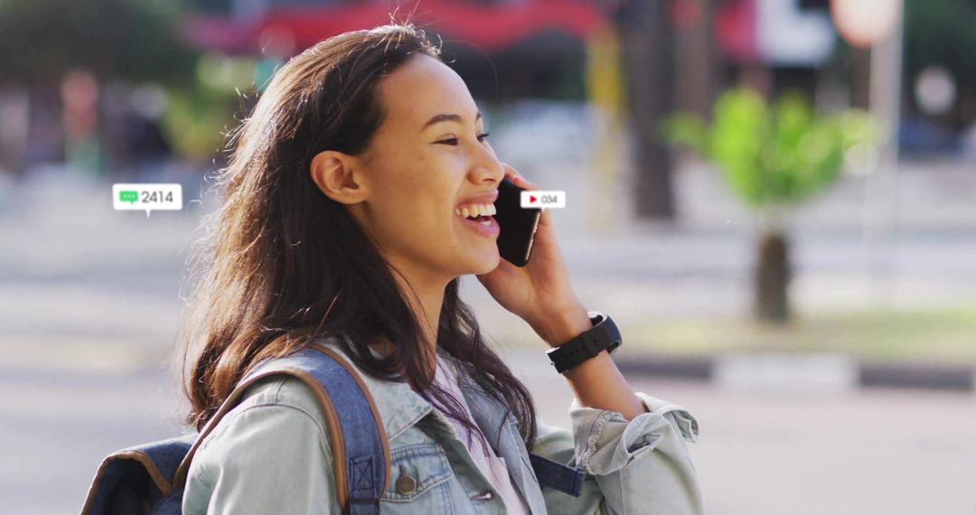 Smiling Asian woman talking on smartphone in denim jacket smartwatch and backpack
