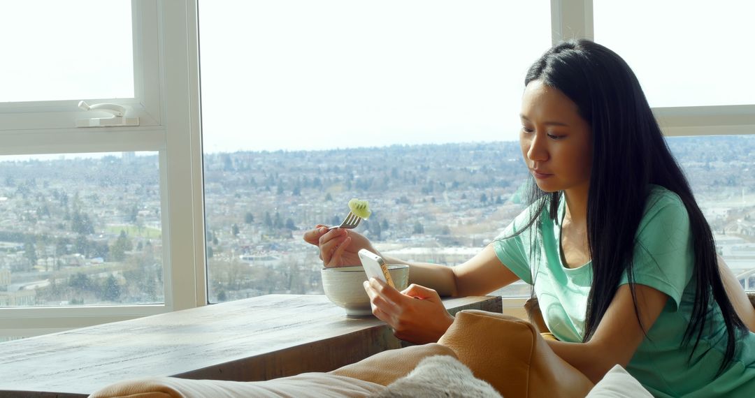 Young woman eating and using smartphone by large window with city view - Free Images, Stock Photos and Pictures on Pikwizard.com