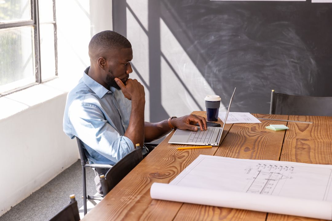 African American Architect Working on Laptop with Blueprints in Light-Filled Workspace
