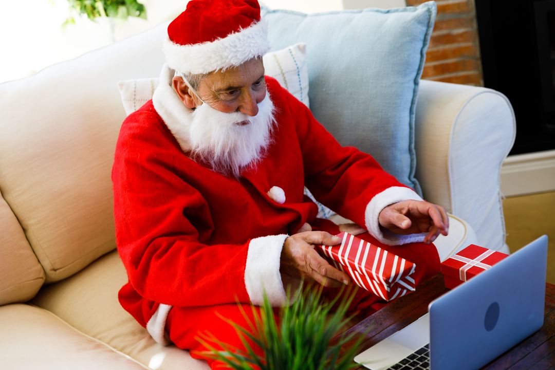Elderly man in Santa costume presents gift on video call, another gift beside laptop. - Free Images, Stock Photos and Pictures on Pikwizard.com