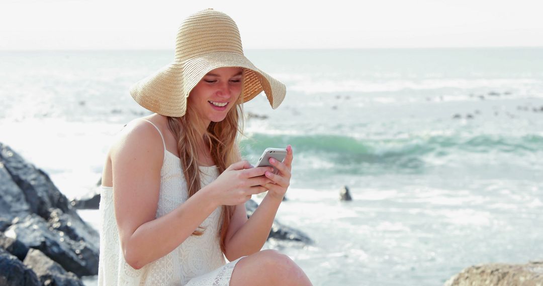 Smiling Woman Wearing Sunhat Using Smartphone by Ocean - Free Images, Stock Photos and Pictures on Pikwizard.com