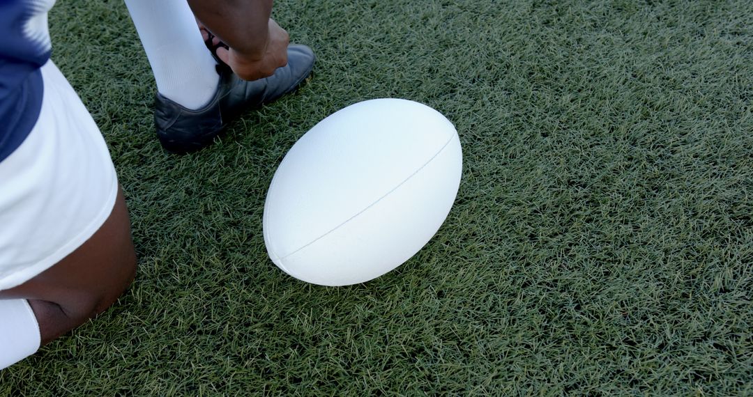 Rugby Player Tying Cleat Beside White Ball on Turf Field