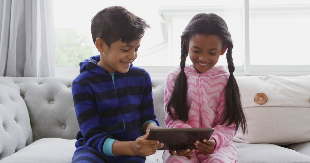 Children in pajamas sharing tablet on sofa smiling - Free Images, Stock Photos and Pictures on Pikwizard.com