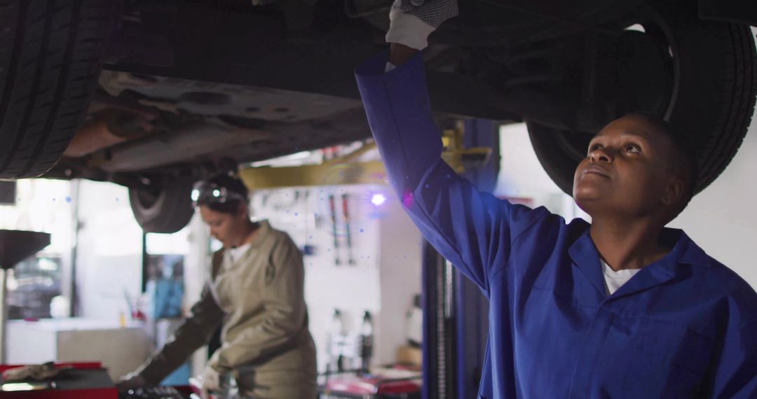 Technician inspecting undercarriage of raised car in garage while colleague checking tools