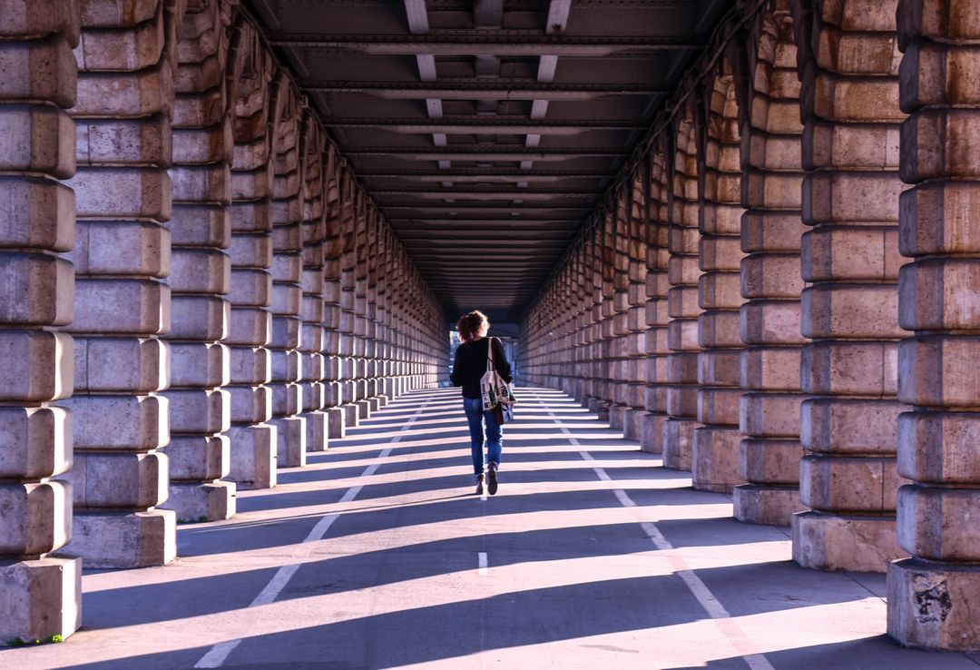 Person Walking in Sunlit Urban Bridge Tunnel with Repeated Columns - Free Images, Stock Photos and Pictures on Pikwizard.com