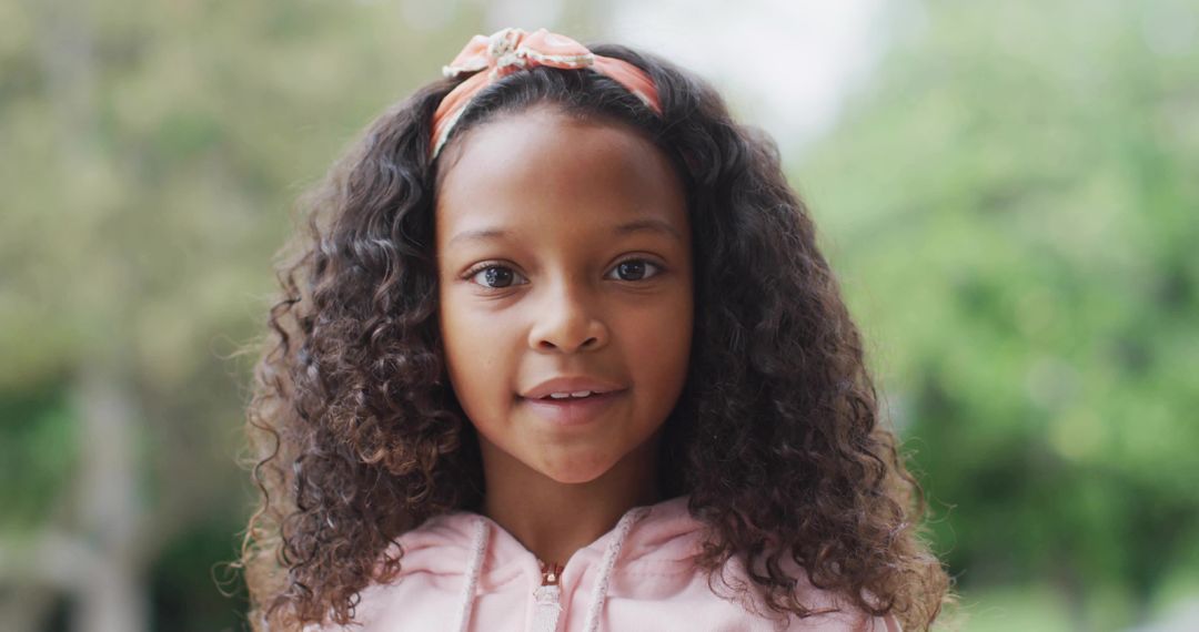 Young girl with curly hair speaking in park wearing pink zip-up hoodie and floral headband