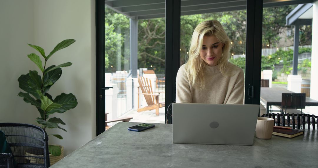 Blonde woman working on laptop at bright minimalist dining table with greenery view