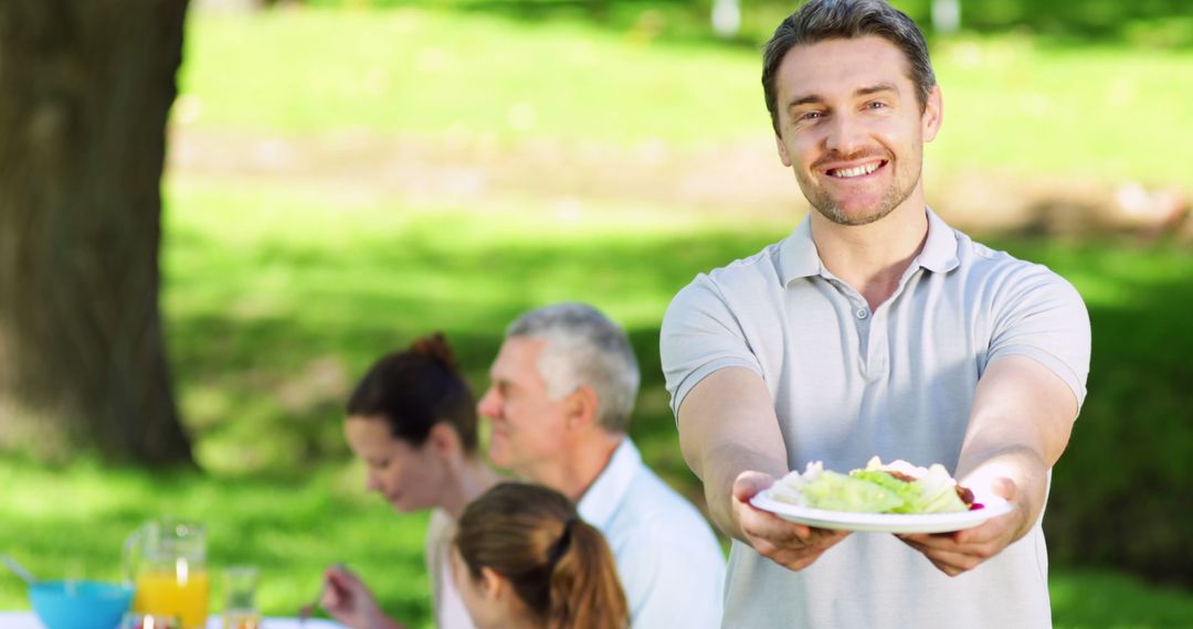 Cheerful Man Showing Plate of Food at Outdoor Family Picnic - Free Images, Stock Photos and Pictures on Pikwizard.com