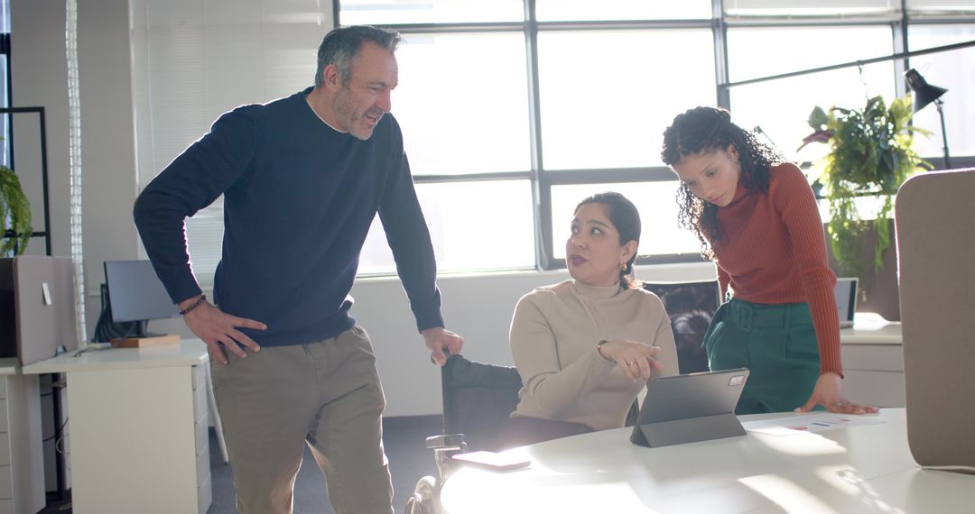 Diverse team collaborating around tablet in sunlit open-plan office, colleague pointing