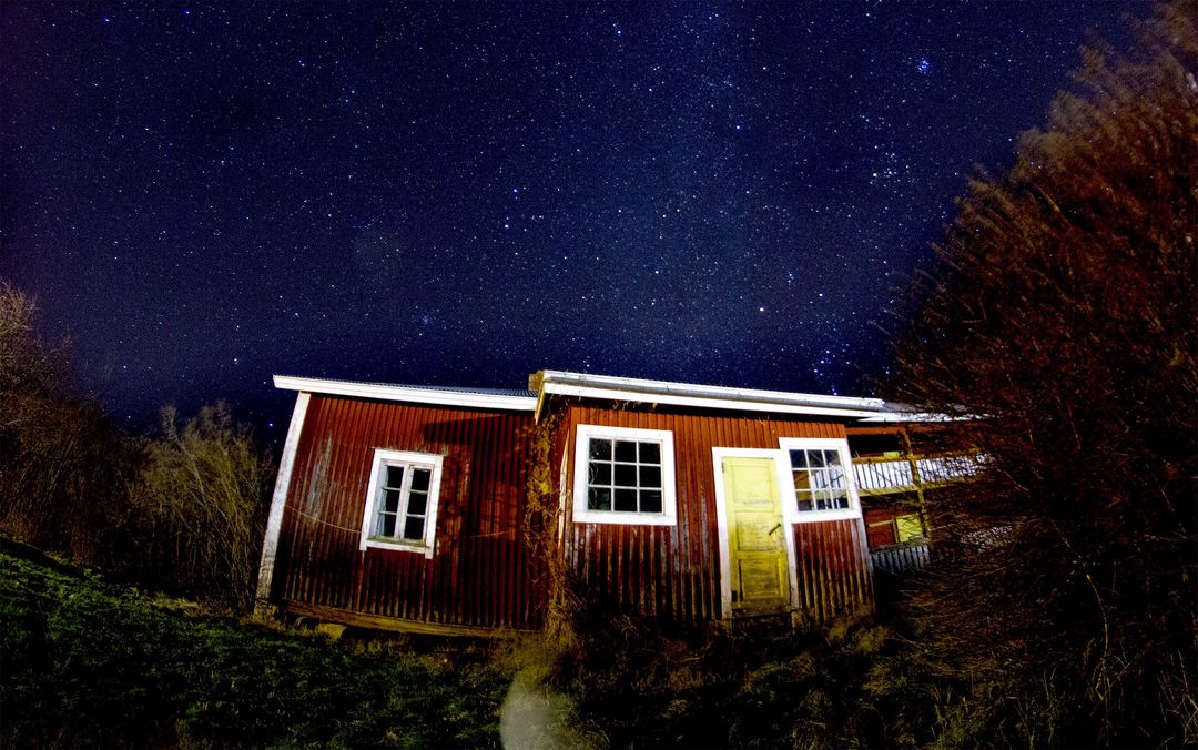 Rustic red cabin standing beneath expansive starry night sky with glowing yellow door