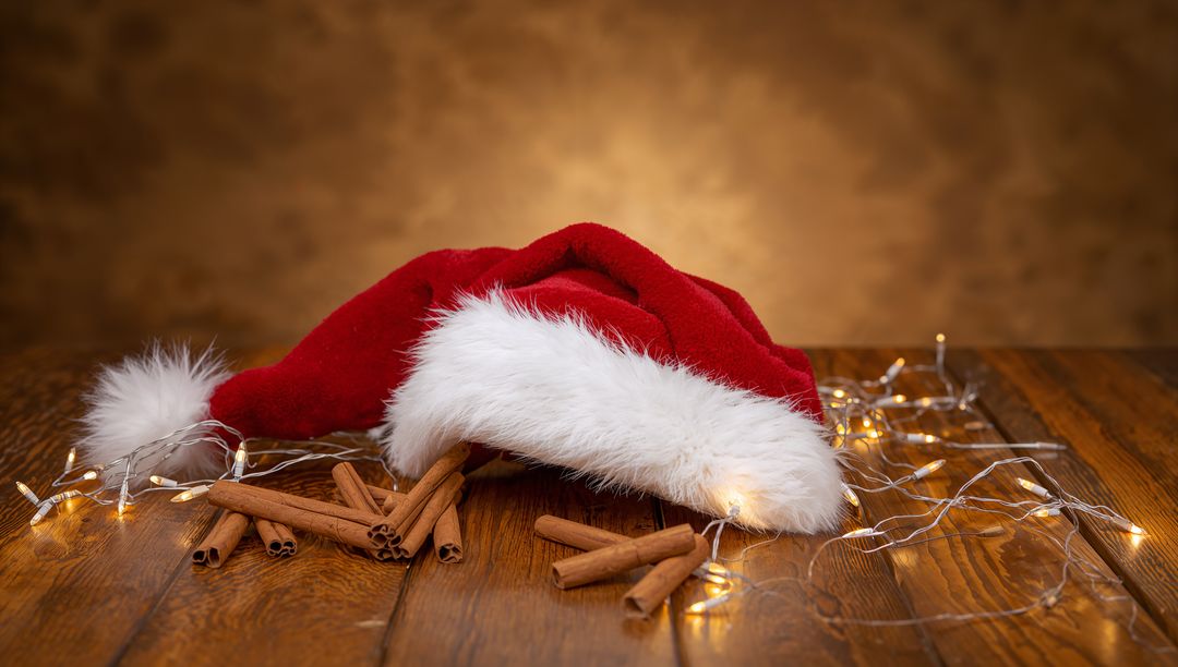 Resting red Santa hat with fur trim on rustic wooden table with cinnamon and fairy lights