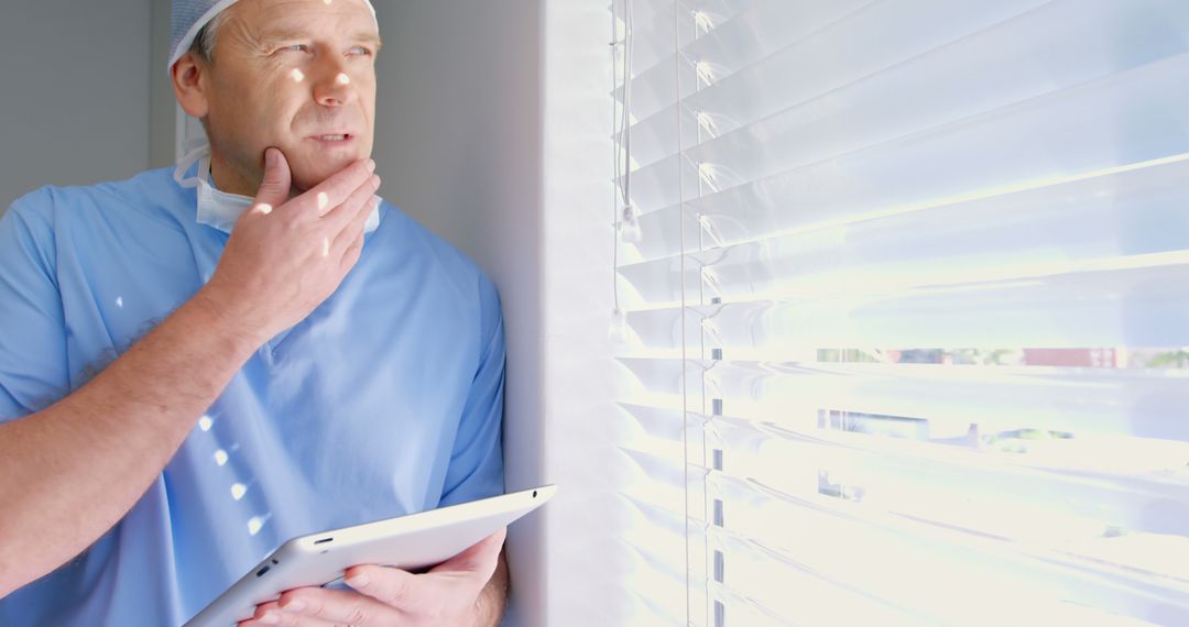 Contemplative Doctor Wearing Surgical Scrubs Holding Tablet Near Window - Free Images, Stock Photos and Pictures on Pikwizard.com