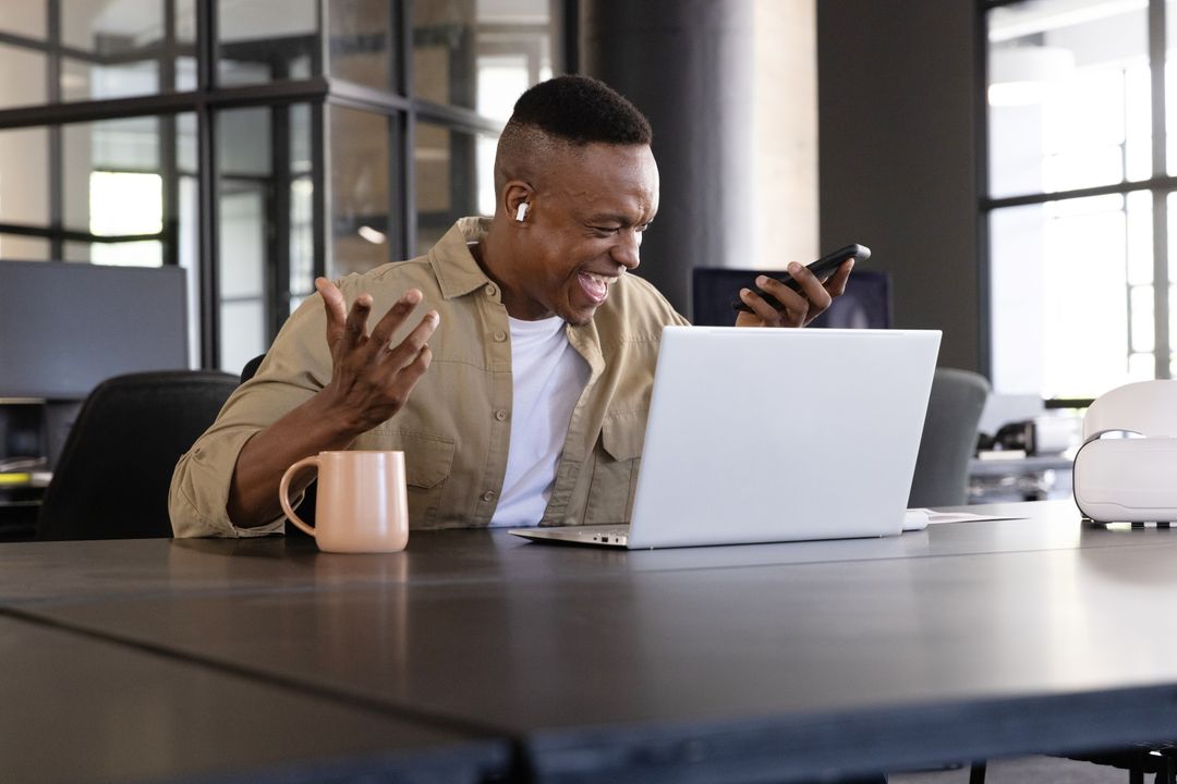 African American man celebrating success on laptop while checking smartphone in office