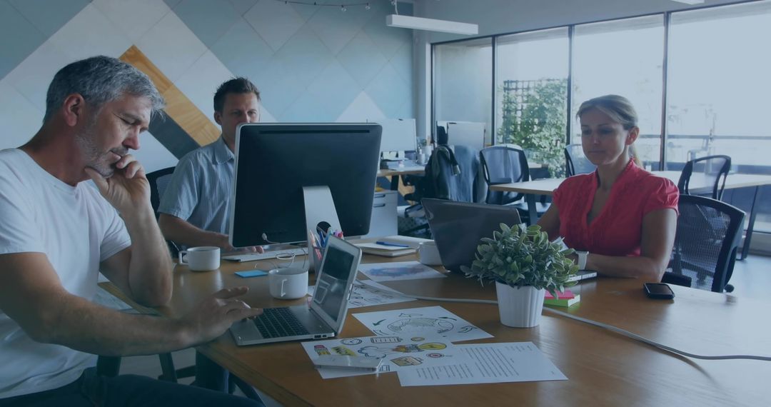 Three coworkers collaborating at modern office table using laptops and reviewing plans